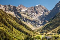 Berglandschaft mit bewaldeten Hängen und schneebedeckten Gipfeln unter blauem Himmel