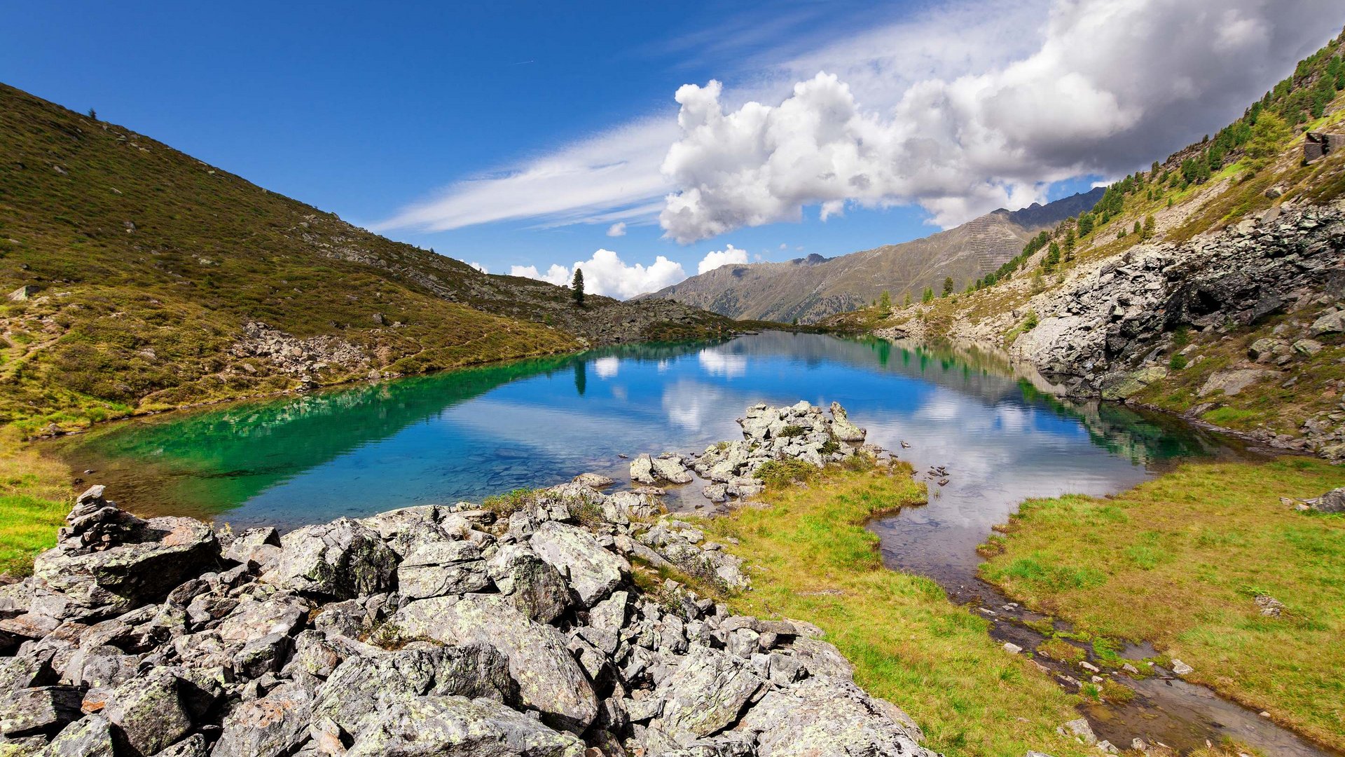 Active holidays at our sports hotel in Pitztal Clear mountain lake water with rocks and green slopes under cloudy blue sky