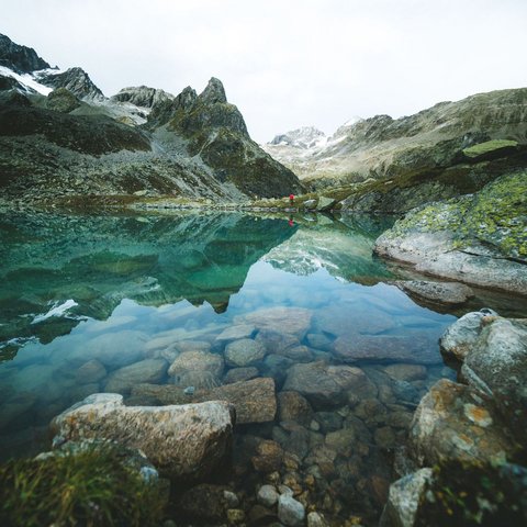 Bilder aus der Sportalm Klares Gebirgssee mit Felsen und Bergspiegelung unter bewölktem Himmel