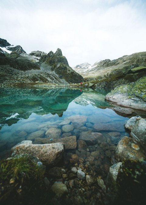 Bilder aus der Sportalm Klares Gebirgssee mit Felsen und Bergspiegelung unter bewölktem Himmel