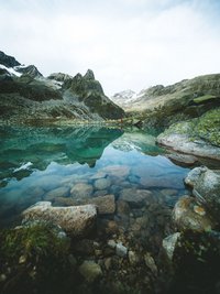 Klares Gebirgssee mit Felsen und Bergspiegelung unter bewölktem Himmel