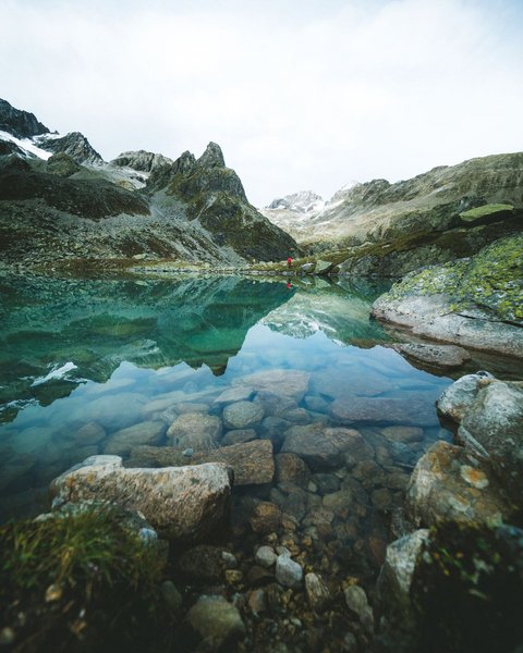 Bilder aus der Sportalm Klares Gebirgssee mit Felsen und Bergspiegelung unter bewölktem Himmel