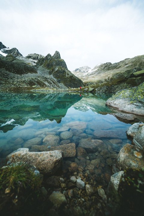 Bilder aus der Sportalm Klares Gebirgssee mit Felsen und Bergspiegelung unter bewölktem Himmel