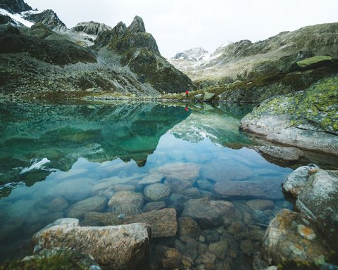 Bilder aus der Sportalm Klares Gebirgssee mit Felsen und Bergspiegelung unter bewölktem Himmel