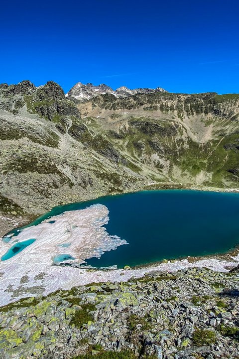 Bilder aus der Sportalm Bergsee mit schmelzendem Schnee und felsigem Gelände unter klarem blauem Himmel