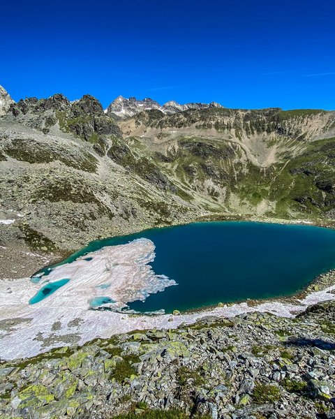 Bilder aus der Sportalm Bergsee mit schmelzendem Schnee und felsigem Gelände unter klarem blauem Himmel