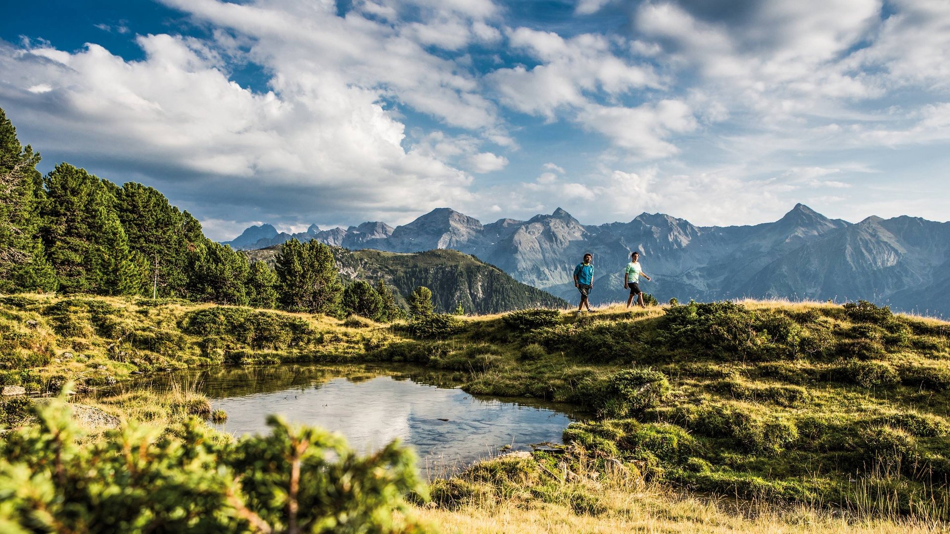 Hiking hotel in Pitztal: nature adventurers, ahoy! Two hikers on a mountain trail with lake and Alps in the background