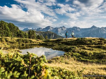 „Nuigkeiten“ aus der Sportalm Zwei Wanderer auf einem Bergweg mit See und Alpen im Hintergrund