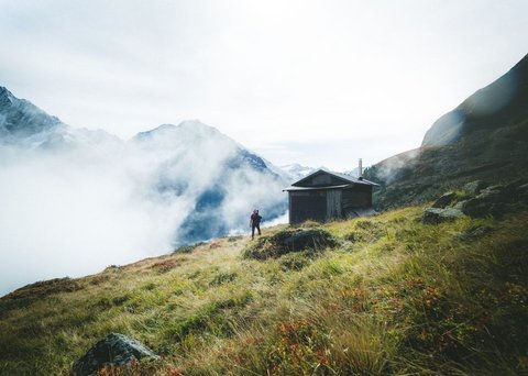 Bilder aus der Sportalm Wanderer auf grasbewachsenem Berg mit Hütte und Nebel in den Alpen