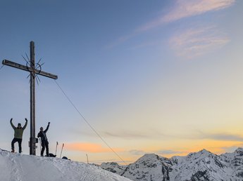 News from Sportalm Two people beside a summit cross on snowy mountains at sunset