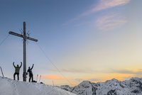 Two people beside a summit cross on snowy mountains at sunset