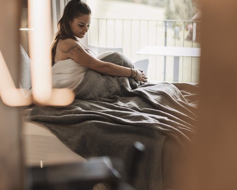 Bilder aus der Sportalm Frau sitzt im Bett mit grauer Decke, Blick aus Fenster auf Wald