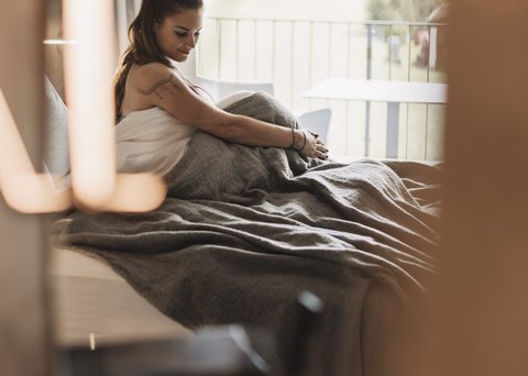 Bilder aus der Sportalm Frau sitzt im Bett mit grauer Decke, Blick aus Fenster auf Wald