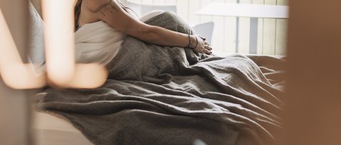 Bilder aus der Sportalm Frau sitzt im Bett mit grauer Decke, Blick aus Fenster auf Wald