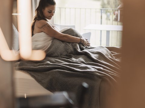 Bilder aus der Sportalm Frau sitzt im Bett mit grauer Decke, Blick aus Fenster auf Wald