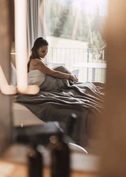 Bilder aus der Sportalm Frau sitzt im Bett mit grauer Decke, Blick aus Fenster auf Wald