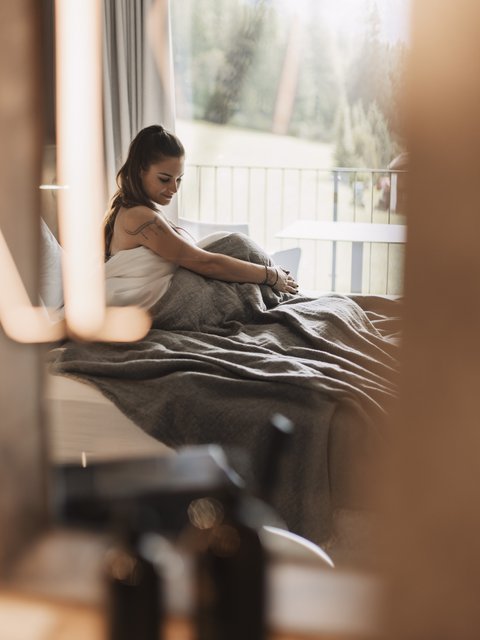 Bilder aus der Sportalm Frau sitzt im Bett mit grauer Decke, Blick aus Fenster auf Wald