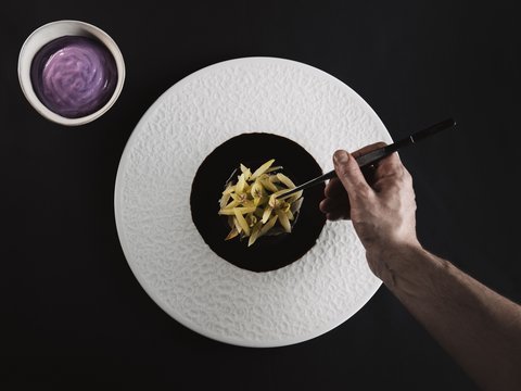 Bilder aus der Sportalm Hand richtet feines Gericht mit Pinzette auf weißem Teller an neben Schüssel mit lila Sauce
