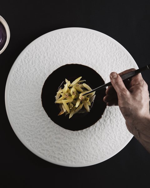 Bilder aus der Sportalm Hand richtet feines Gericht mit Pinzette auf weißem Teller an neben Schüssel mit lila Sauce