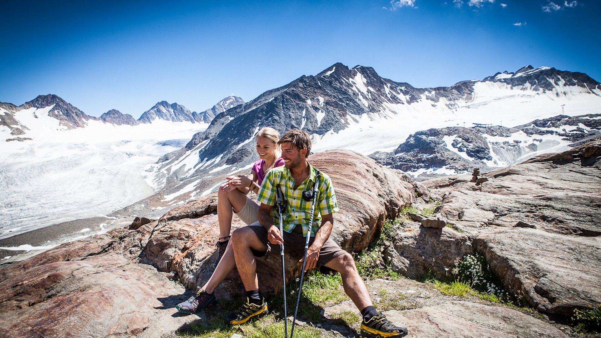 Hiking hotel in Pitztal: nature adventurers, ahoy! Couple resting on rocks while hiking with glacier and mountains in the background