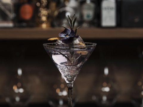 Unser Restaurant im Pitztal Cocktailglas mit klarem Drink, Eiswürfel und essbarer Blume vor Bar mit Flaschen