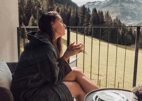 Bilder aus der Sportalm Frau in Bademantel sitzt auf Balkon mit Bergblick am Tisch