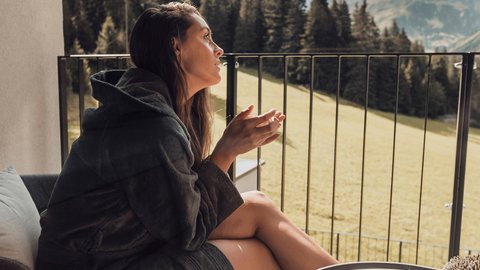 Bilder aus der Sportalm Frau in Bademantel sitzt auf Balkon mit Bergblick am Tisch
