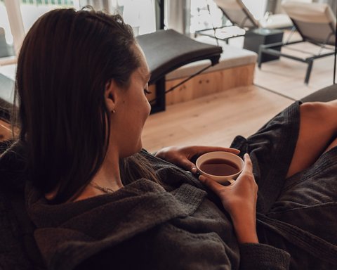 Bilder aus der Sportalm Frau entspannt in Bademantel und trinkt Tee in modernem Spa mit Aussicht