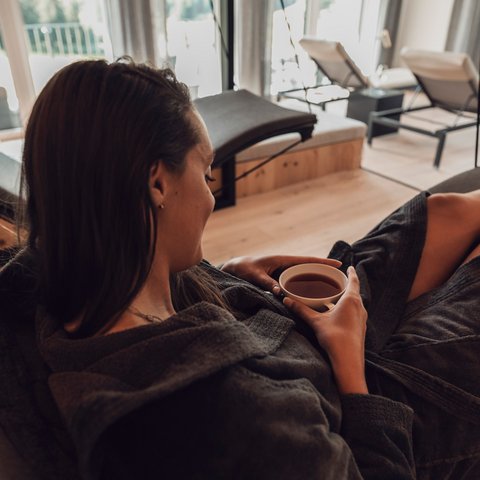 Bilder aus der Sportalm Frau entspannt in Bademantel und trinkt Tee in modernem Spa mit Aussicht