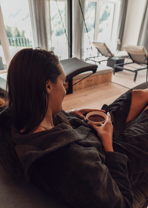 Bilder aus der Sportalm Frau entspannt in Bademantel und trinkt Tee in modernem Spa mit Aussicht