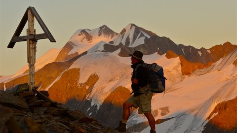 Bilder aus der Sportalm Wanderer mit Rucksack auf Bergpfad neben Holzkreuz bei Sonnenuntergang