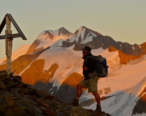 Bilder aus der Sportalm Wanderer mit Rucksack auf Bergpfad neben Holzkreuz bei Sonnenuntergang