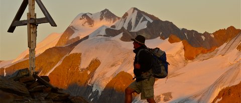 Bilder aus der Sportalm Wanderer mit Rucksack auf Bergpfad neben Holzkreuz bei Sonnenuntergang