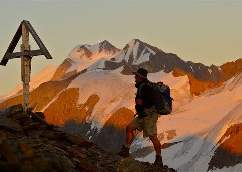 Bilder aus der Sportalm Wanderer mit Rucksack auf Bergpfad neben Holzkreuz bei Sonnenuntergang