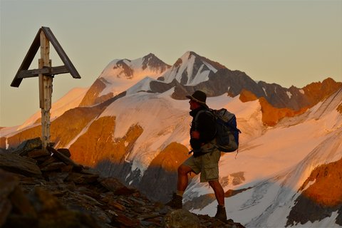 Bilder aus der Sportalm Wanderer mit Rucksack auf Bergpfad neben Holzkreuz bei Sonnenuntergang