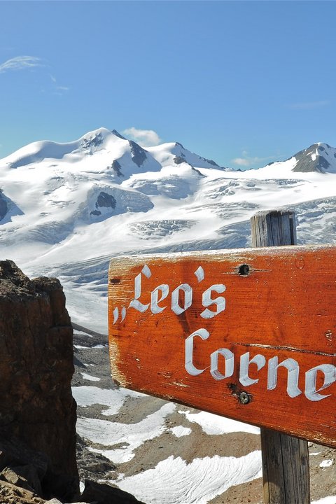 Bilder aus der Sportalm Holzschild Leo's Corner vor schneebedeckten Bergen und blauem Himmel