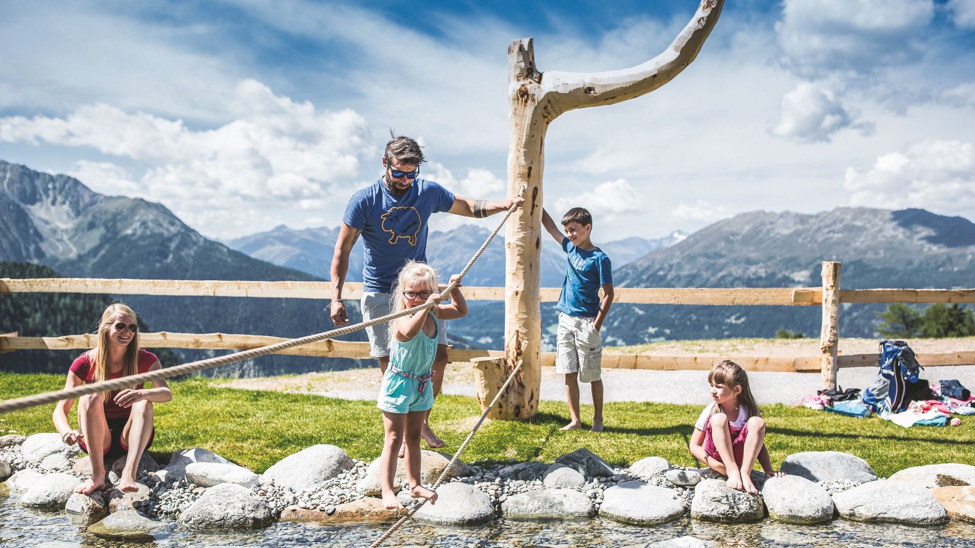 Hiking hotel in Pitztal: nature adventurers, ahoy! Family playing barefoot by mountain stream with rope and stones in sunshine