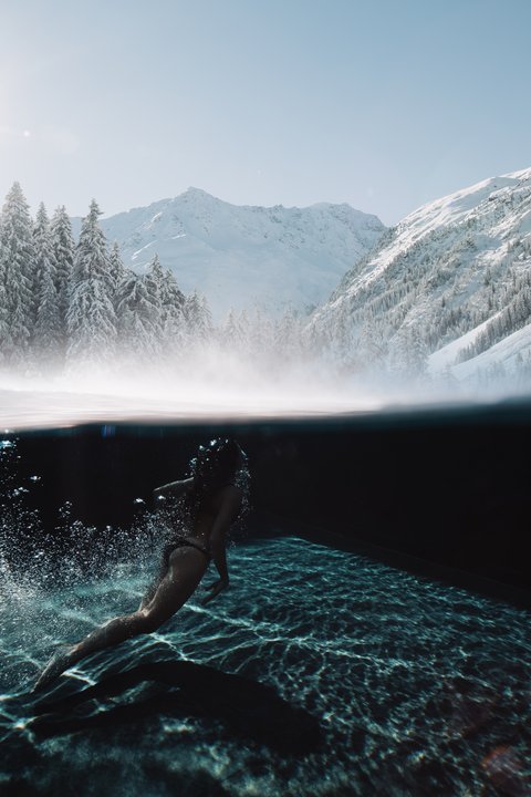 Bilder aus der Sportalm Frau schwimmt im klaren Wasser mit verschneiten Bergen und Sonnenlicht im Hintergrund