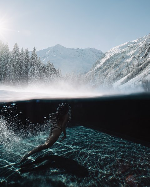 Bilder aus der Sportalm Frau schwimmt im klaren Wasser mit verschneiten Bergen und Sonnenlicht im Hintergrund