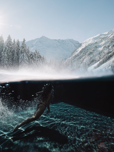 Bilder aus der Sportalm Frau schwimmt im klaren Wasser mit verschneiten Bergen und Sonnenlicht im Hintergrund
