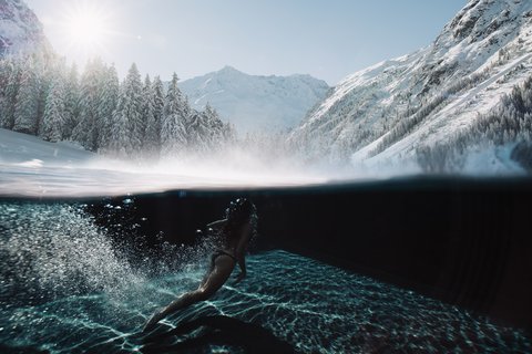 Bilder aus der Sportalm Frau schwimmt im klaren Wasser mit verschneiten Bergen und Sonnenlicht im Hintergrund