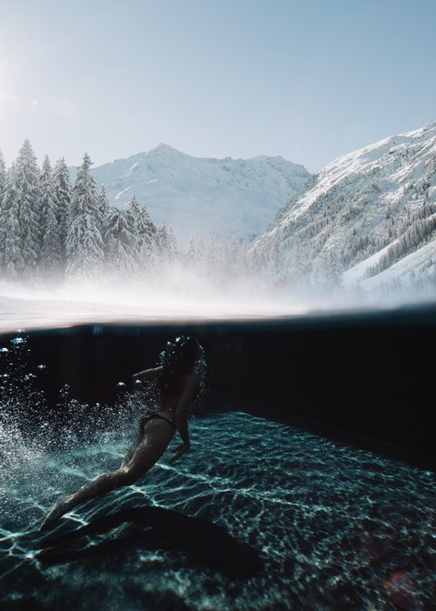Bilder aus der Sportalm Frau schwimmt im klaren Wasser mit verschneiten Bergen und Sonnenlicht im Hintergrund