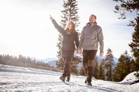 Paar geht im Schnee spazieren, Frau zeigt nach oben, Berge und Bäume im Hintergrund