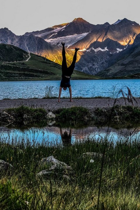 Bilder aus der Sportalm Person macht Handstand vor Bergsee mit Sonnenlicht auf Bergen