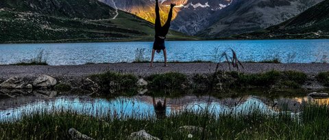Bilder aus der Sportalm Person macht Handstand vor Bergsee mit Sonnenlicht auf Bergen