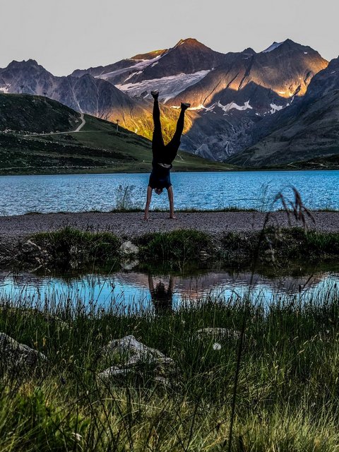Pictures from Sportalm Person doing handstand by mountain lake with sunlight on peaks