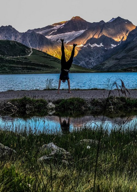 Bilder aus der Sportalm Person macht Handstand vor Bergsee mit Sonnenlicht auf Bergen