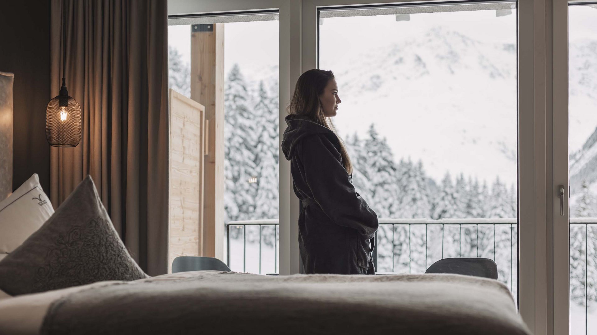 Energiereserven aufladen in der Sportalm Frau im Bademantel blickt aus Hotelzimmer auf verschneite Berge