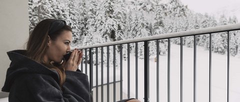 Bilder aus der Sportalm Frau im Bademantel trinkt Heißgetränk auf Balkon mit verschneitem Bergblick