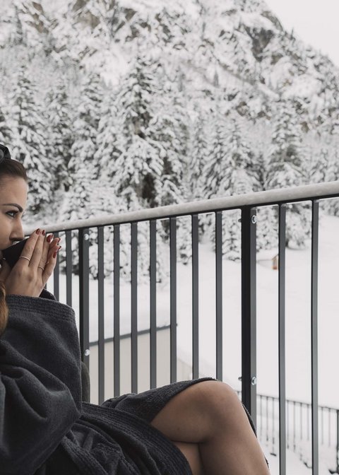 Bilder aus der Sportalm Frau im Bademantel trinkt Heißgetränk auf Balkon mit verschneitem Bergblick